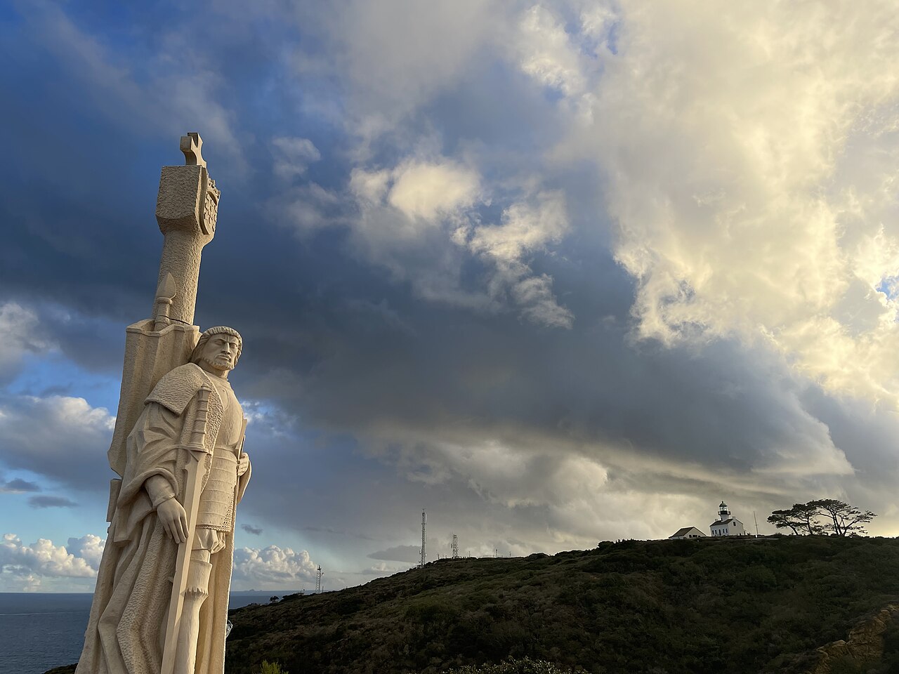 The limestone statue of Juan Rodríguez Cabrillo in the foreground, with the white Old Point Loma Lighthouse on the hill behind it and the Pacific Ocean to the left.
